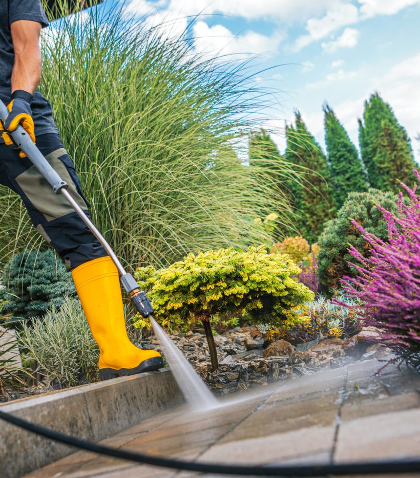 A person in yellow boots operates a pressure washer, clearing dirt and debris from a garden path lined with colorful shrubs and greenery.