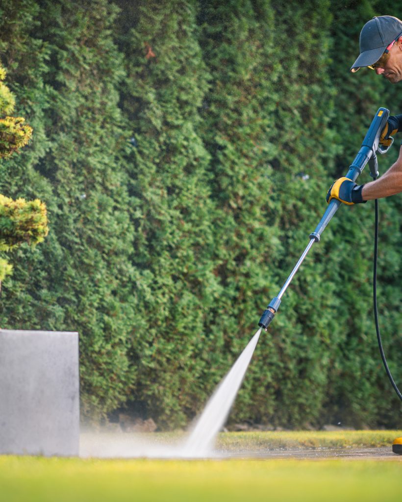 A landscaper, wearing yellow boots and gloves, is cleaning paved surfaces with a pressure washer in a vibrant garden surrounded by tall hedges.